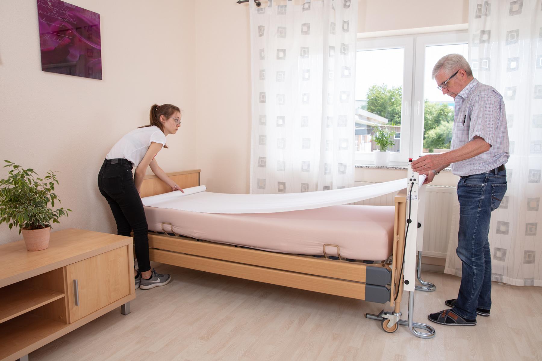 Das pulla Bettlaken wird über die Liegefläche eines Pflegebetts gezogen.